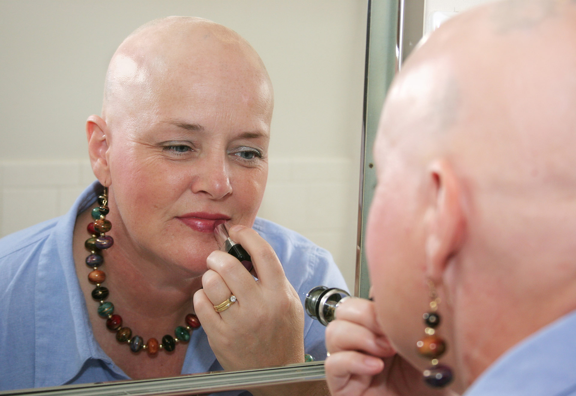 A woman bald from a health problem putting on makeup in the mirror.